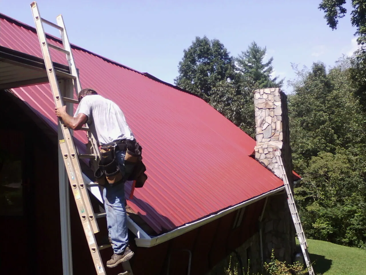 Roofer on a ladder beside a red metal roof and stone chimney — Soffit & Fascia Repair by Roofing Brothers in Frederica